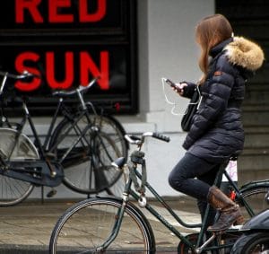 Jeune fille arrêtée avec son vélo sur le bord de la chaussée en train de consulter son smartphone et portant des écouteurs blancs (photo de "27707" - domaine public)